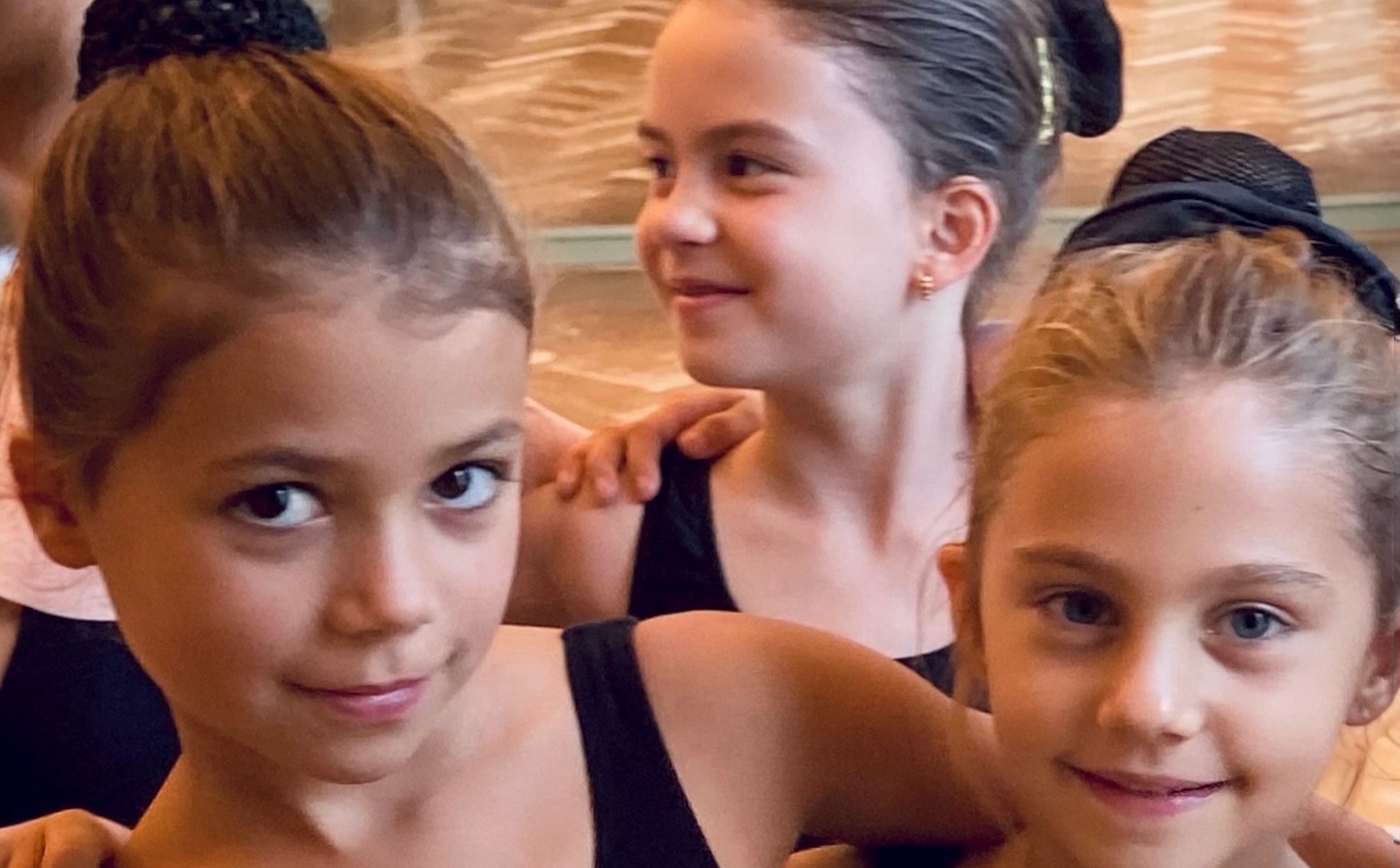 Children from Mirella Simoncini’s Junior Ballet class in The Hague, one smiling at the camera with an arm around a classmate, while another dancer in the background looks up and laughs.