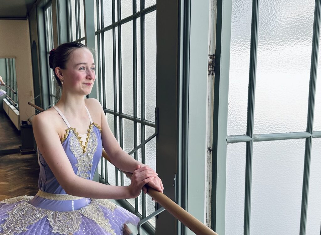 Ballet student in tutu leans dreamily on the barre, smiling and looking out the window, something good is coming.