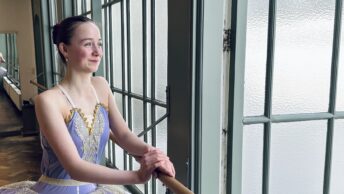 Ballet student in tutu leans dreamily on the barre, smiling and looking out the window, something good is coming.