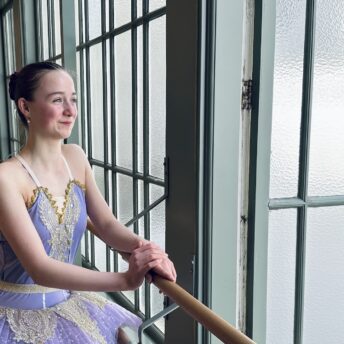 Ballet student in tutu leans dreamily on the barre, smiling and looking out the window, something good is coming.