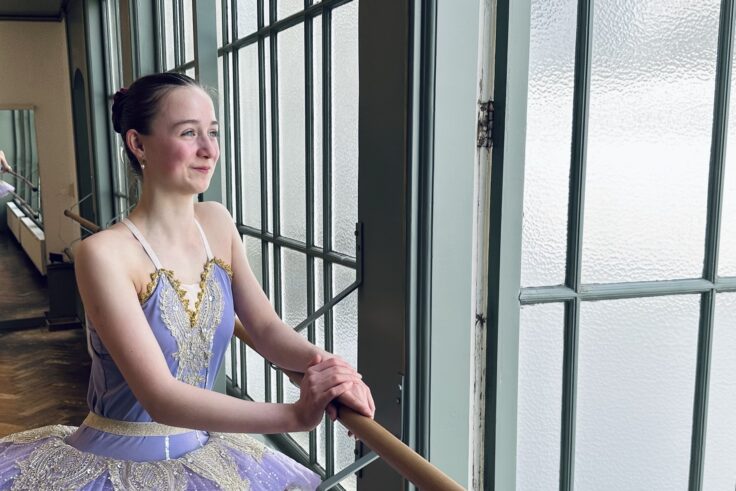 Ballet student in tutu leans dreamily on the barre, smiling and looking out the window, something good is coming.